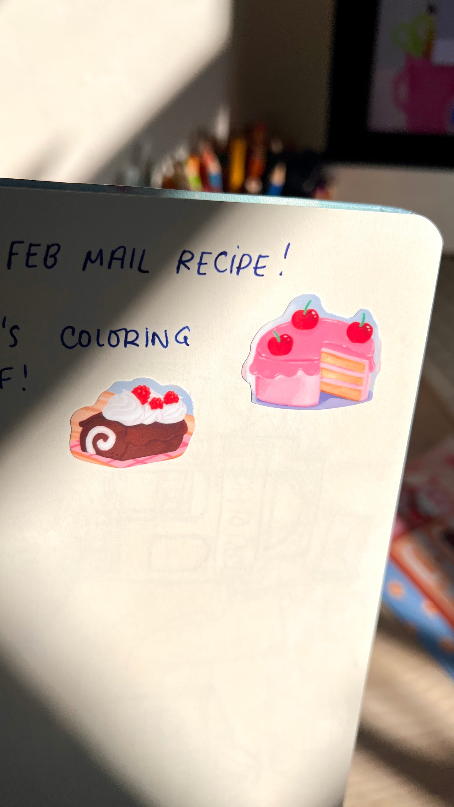 Notebook with stickers of cakes and text about a 'Feb Mail Recipe' and coloring.