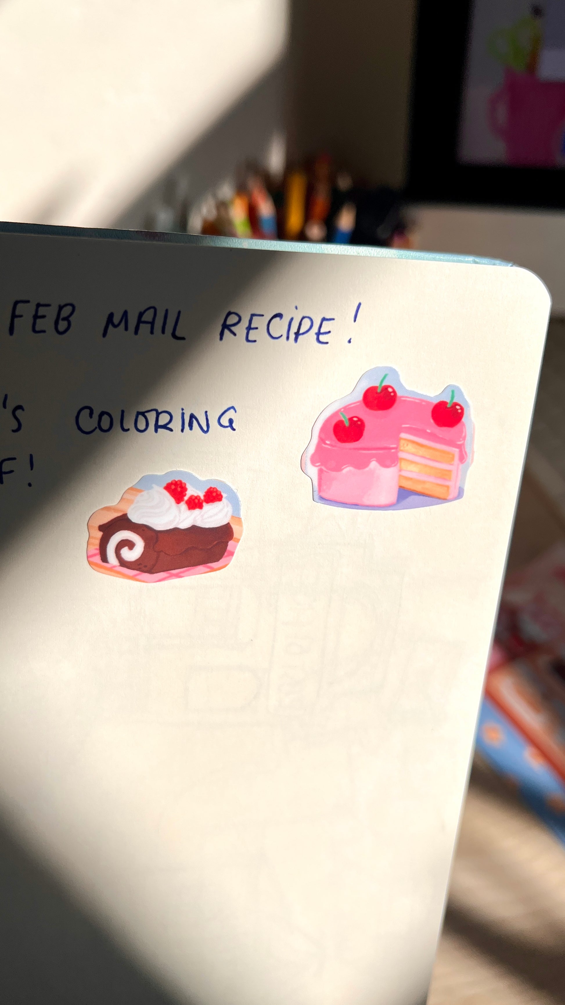 Notebook with stickers of cakes and text about a 'Feb Mail Recipe' and coloring.