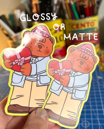 Two cartoon bear stickers with a heart and gift, held by a hand, with text 'Glossy or Matte' above.