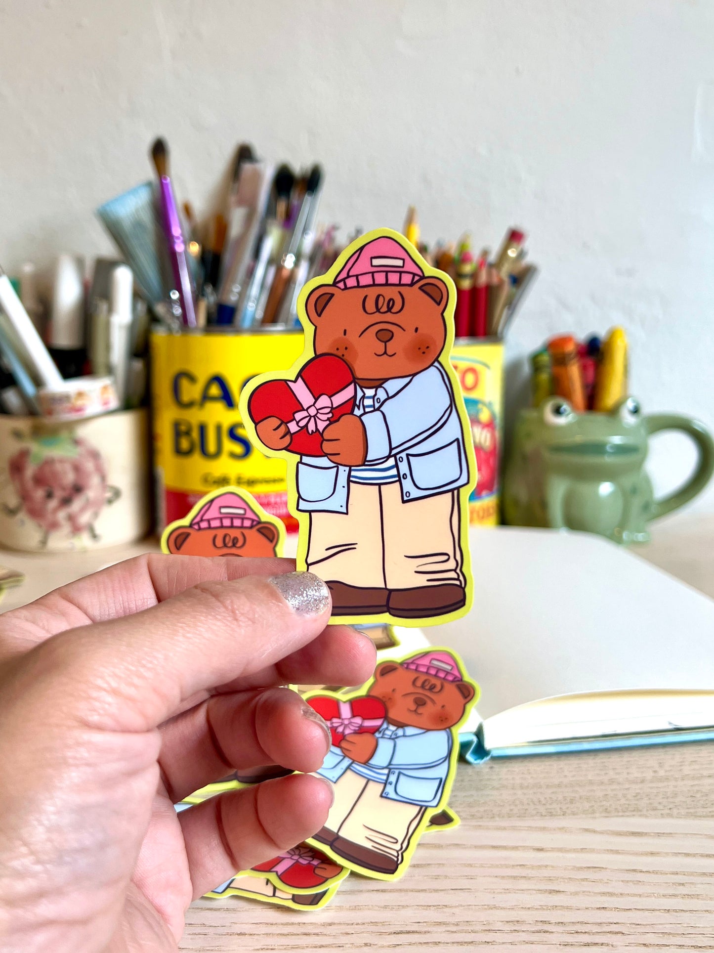 Hand holding a sticker of a bear character with a heart, with a desk and stationery items in the background.