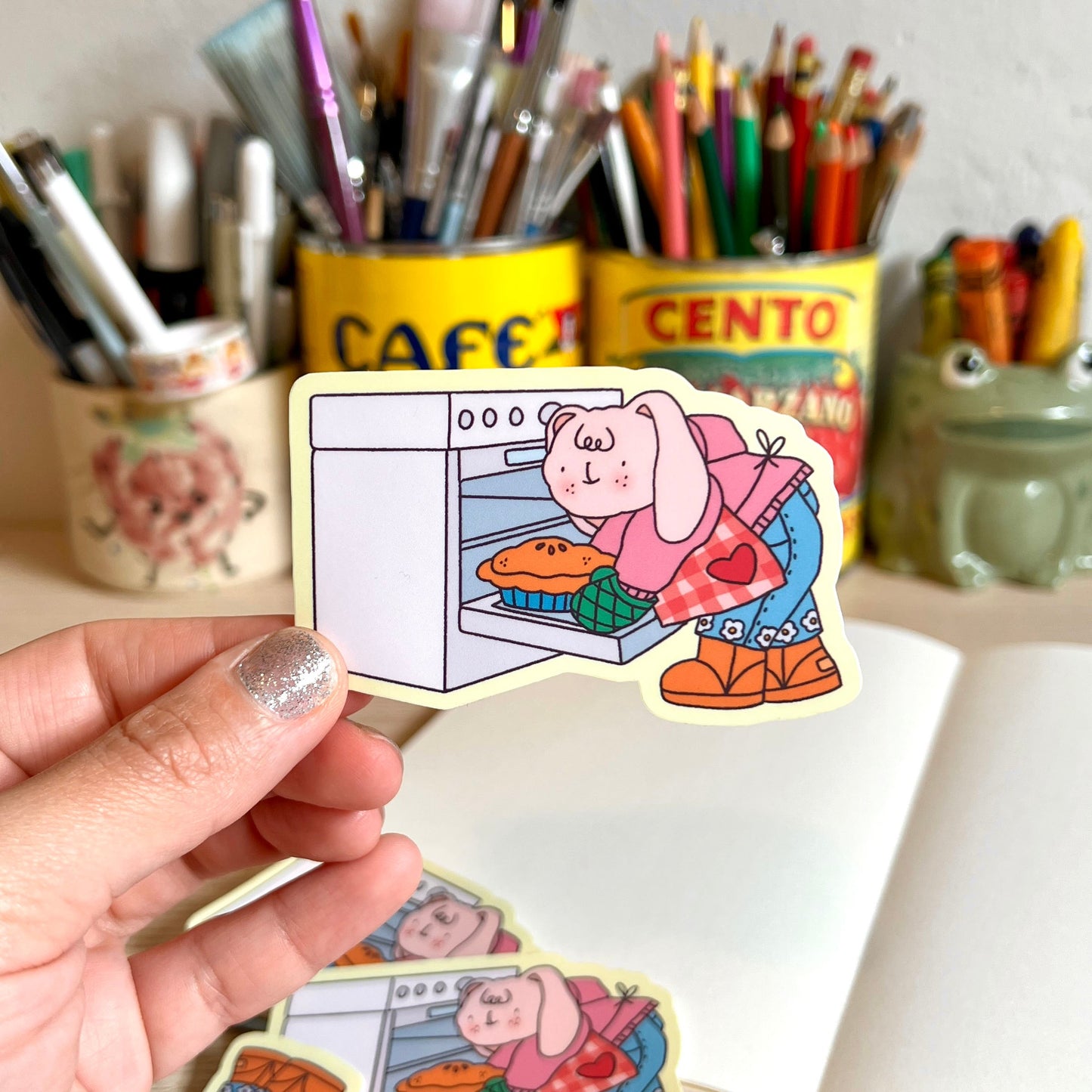 Hand holding a sticker of a cartoon character baking a pie with stationery items in the background.