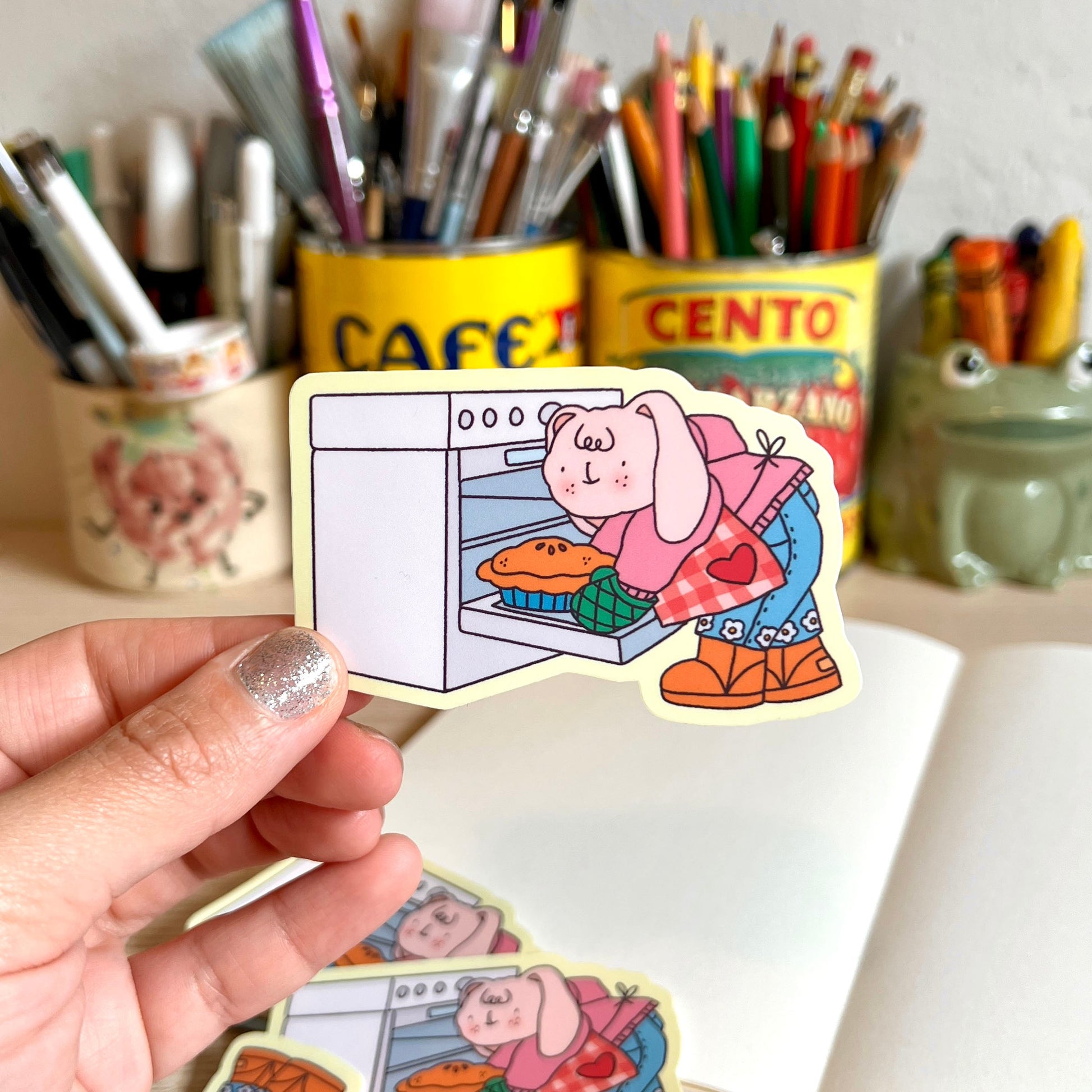 Hand holding a sticker of a cartoon character baking a pie with stationery items in the background.