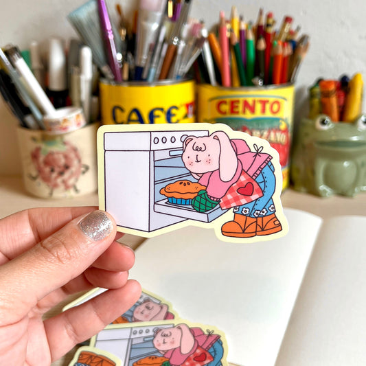 Hand holding a sticker of a cartoon character baking a pie with stationery items in the background.