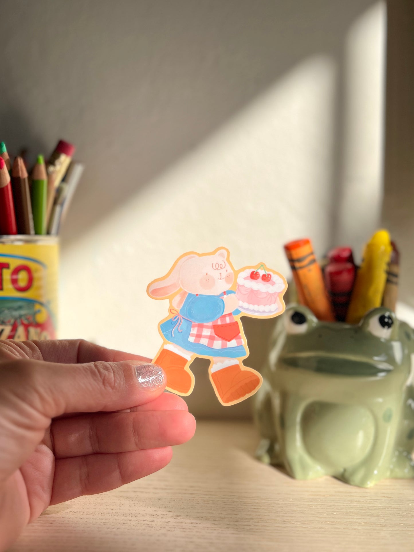 A hand holding a sticker featuring a cute cartoon bunny holding a pink cake