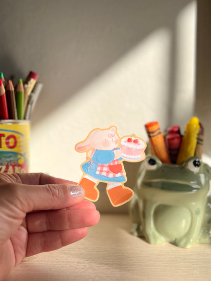 A hand holding a sticker featuring a cute cartoon bunny holding a pink cake